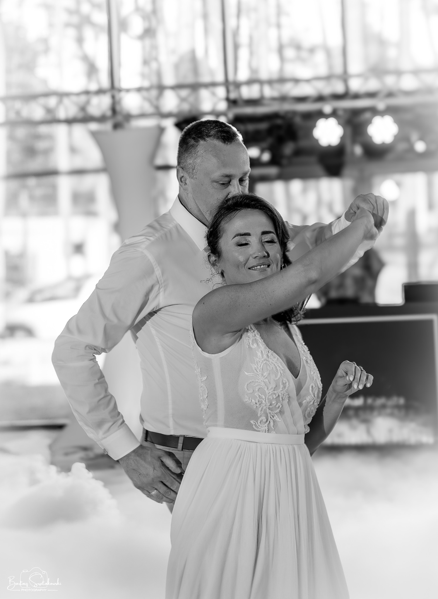 Czarno-białe zdjęcie pary młodej w tańcu, on w koszuli, ona w sukni z koronkową górą, oboje uśmiechnięci na tle dekoracji ślubnej i sztucznego dymu.