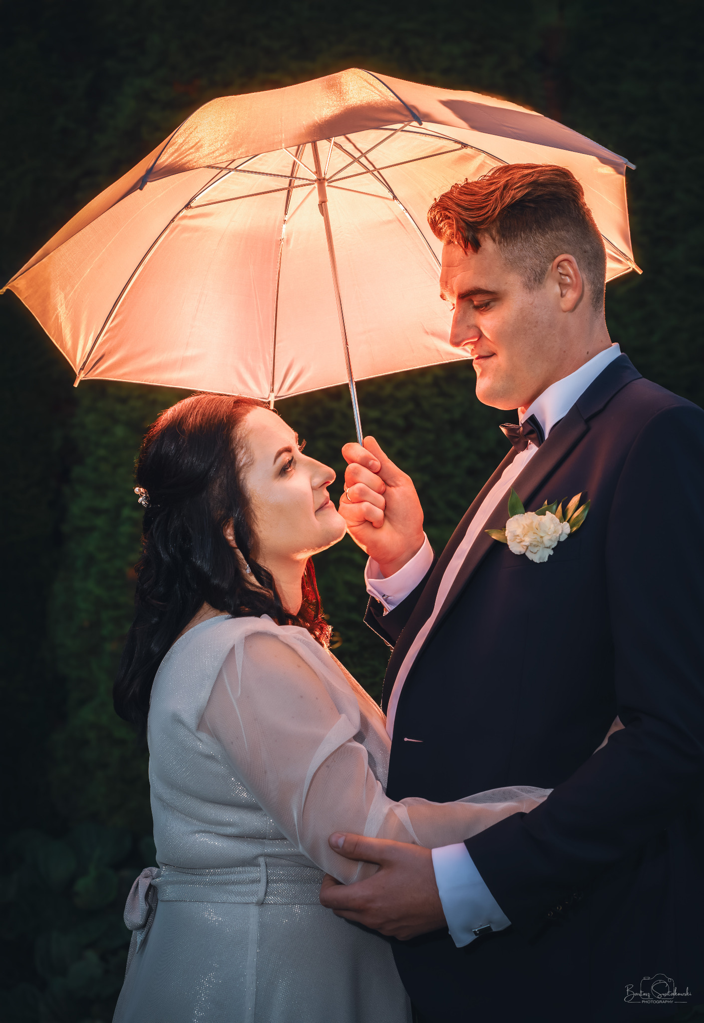 Para młoda w objęciach pod parasolem, oświetlona ciepłym światłem. Uśmiechnięci, w eleganckich strojach, z ciemnozielonym tłem. Delikatne światło podkreśla detale.