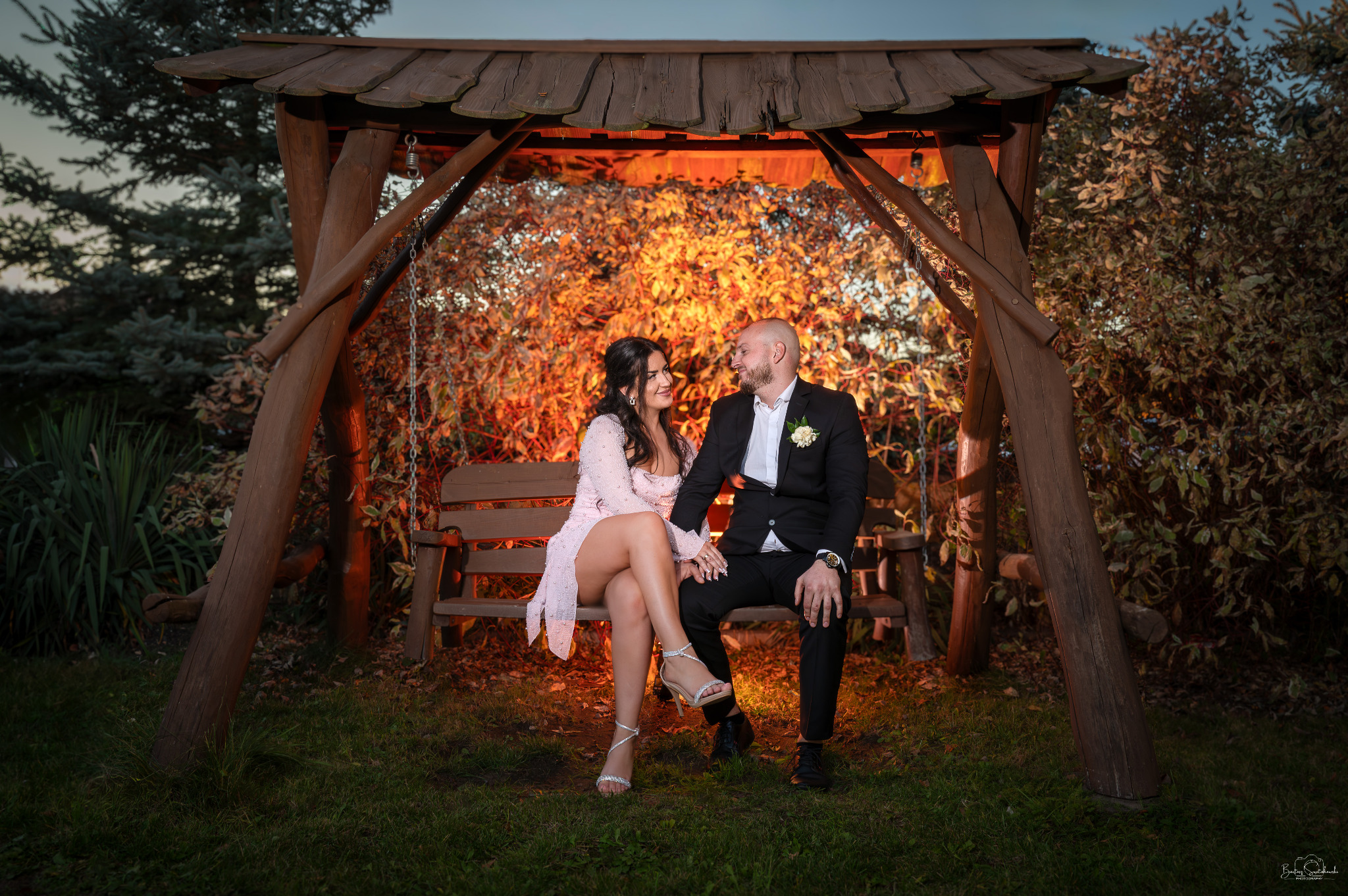 Zakochana para siedzi na drewnianej huśtawce w ogrodzie, oświetlona ciepłym światłem. Kobieta w sukience, mężczyzna w garniturze, patrzą na siebie z uśmiechem.