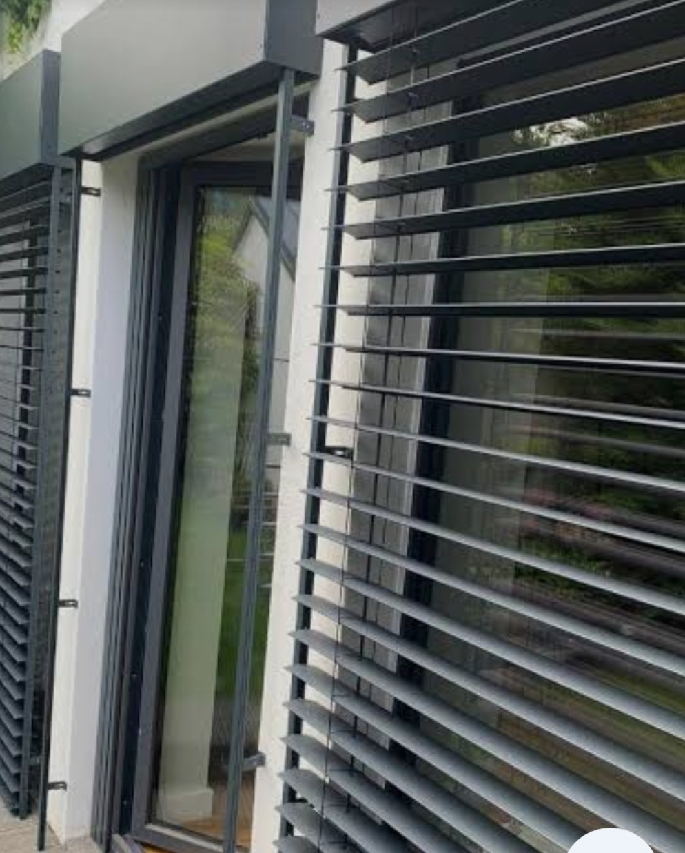 Nowoczesne, szare żaluzje fasadowe zamontowane na oknie budynku. Widoczne detale konstrukcji i odbicie otoczenia w szybie.