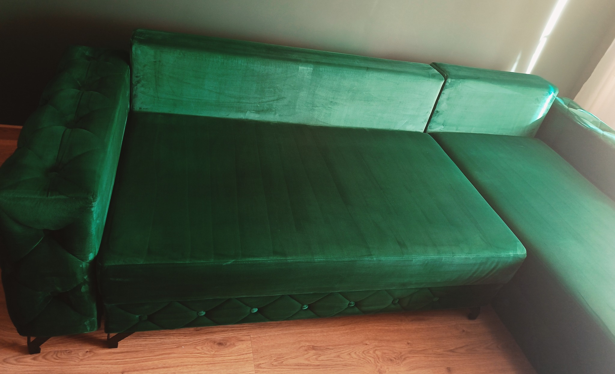 Szmaragdowa, welurowa kanapa narożna z pikowanym wykończeniem, ustawiona na drewnianej podłodze. Widoczne subtelne załamania światła na tkaninie.