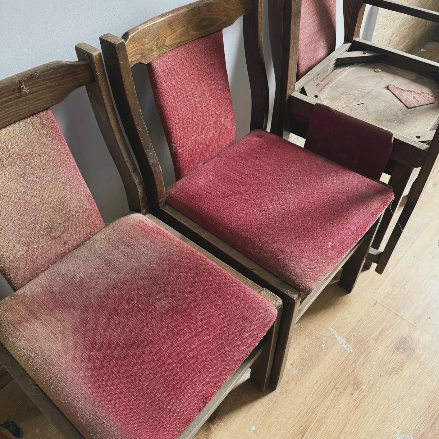 Stare, drewniane krzesła z czerwonymi, mocno zabrudzonymi siedziskami, ustawione obok siebie na podłodze. Widoczne ślady użytkowania i plamy na tapicerce.