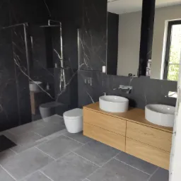Nowoczesna łazienka z czarnymi płytkami imitującymi marmur, dwiema okrągłymi umywalkami na drewnianej szafce, kabiną prysznicową typu walk-in i szarą podłogą z płytek.
