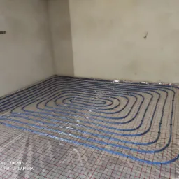 Niebieskie rury ogrzewania podłogowego ułożone spiralnie na siatce w surowym pomieszczeniu przed wylewką.