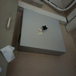 AMT Karotel s.c. - Szara skrzynka telekomunikacyjna z czarnym pokrętłem, przymocowana do ściany, widoczne kable w białej osłonie i naklejka z informacjami o produkcie.