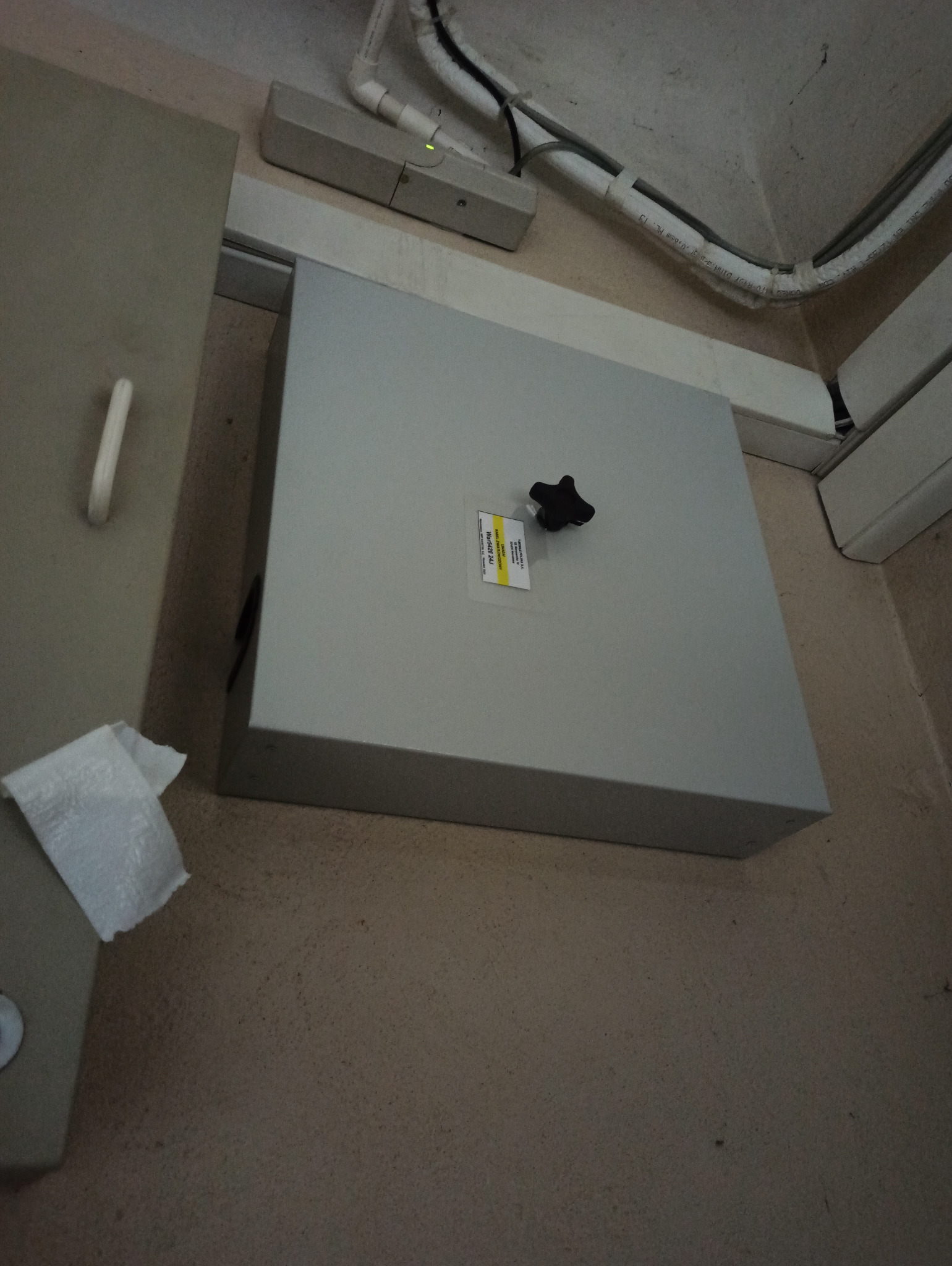 Szara skrzynka telekomunikacyjna z czarnym pokrętłem, przymocowana do ściany, widoczne kable w białej osłonie i naklejka z informacjami o produkcie.