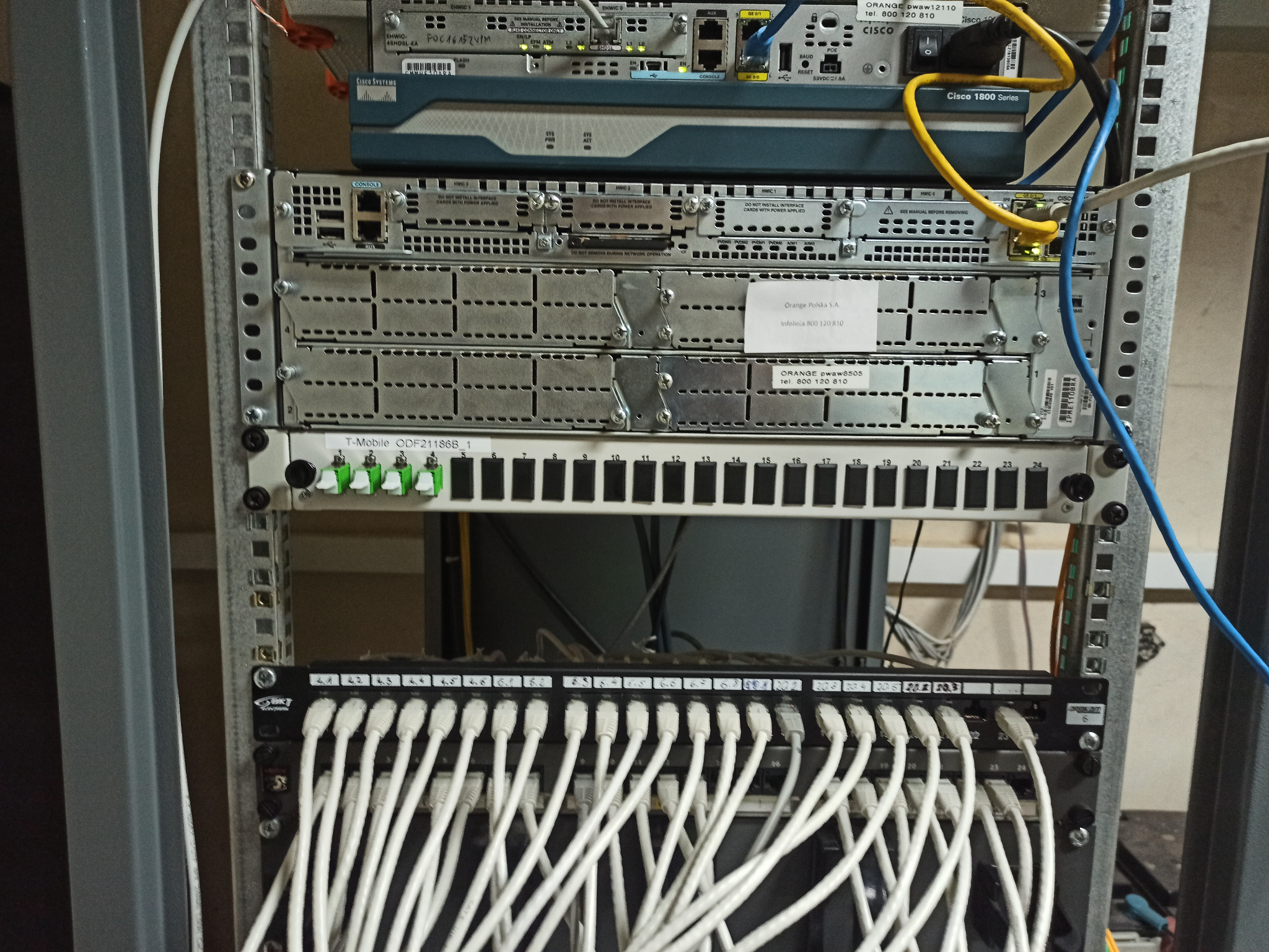 Szafa rack z zamontowanymi urządzeniami sieciowymi, w tym routerami Cisco i patch panelem z podłączonymi kablami Ethernet. Widoczne etykiety Orange Polska i T-Mobile na urządzeniach.