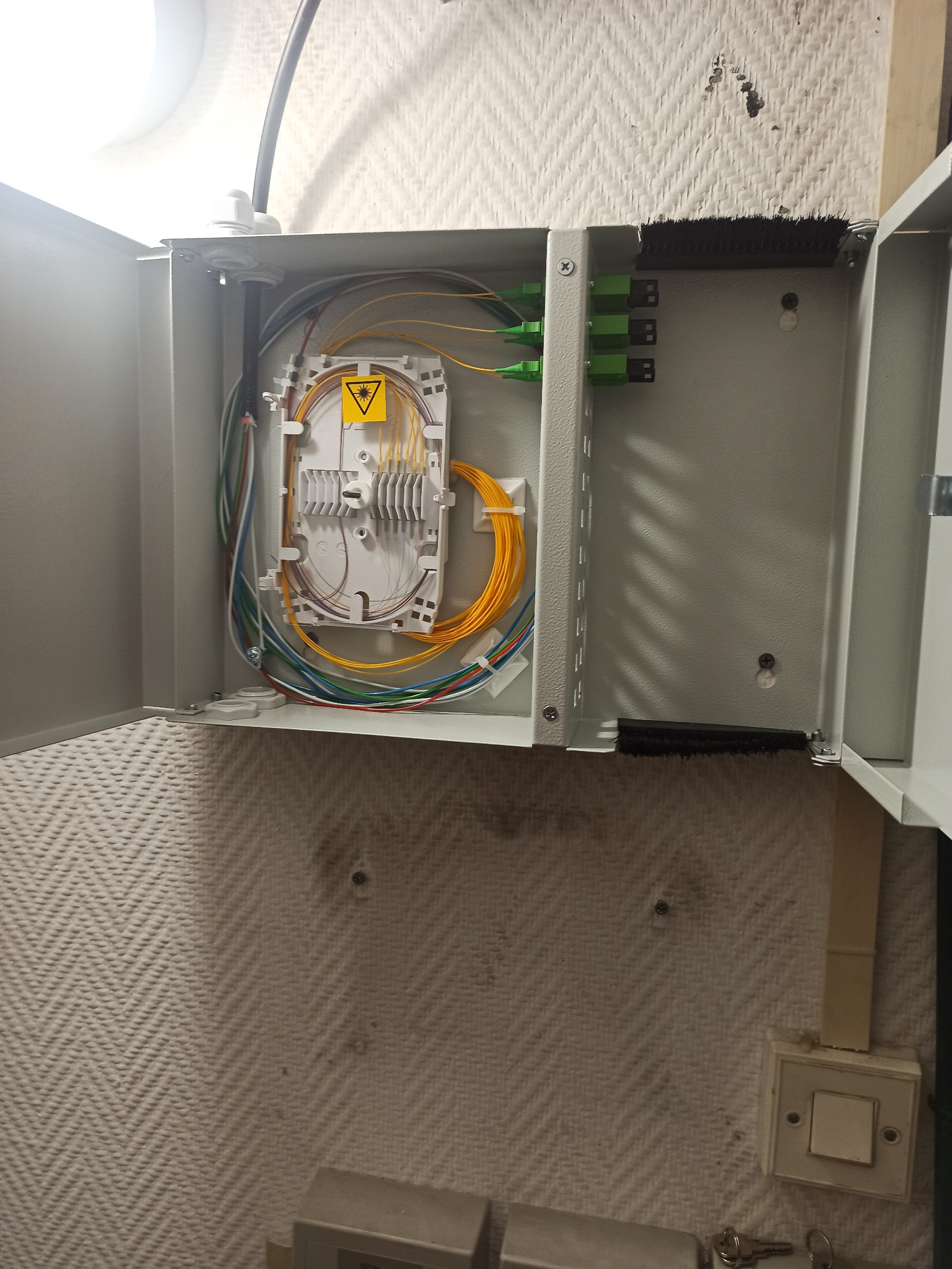 Otwarta skrzynka telekomunikacyjna na ścianie z widocznym spliterem światłowodowym, kable światłowodowe w różnych kolorach oraz złączki światłowodowe.