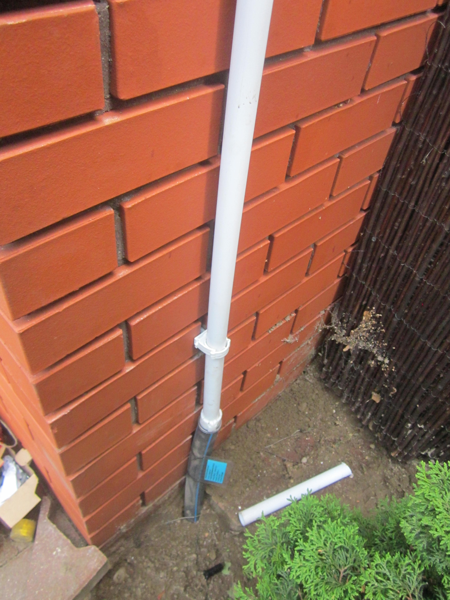Biała rura telekomunikacyjna przymocowana do ceglanej ściany zewnętrznej budynku, z widocznym fragmentem instalacji w ziemi i luźnym odcinkiem rury obok krzewu.