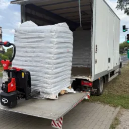 Rozładunek pelletu z ciężarówki za pomocą wózka paletowego, widoczny znak drogowy z kierunkami na Wągrowiec i Poznań. Dzień.
