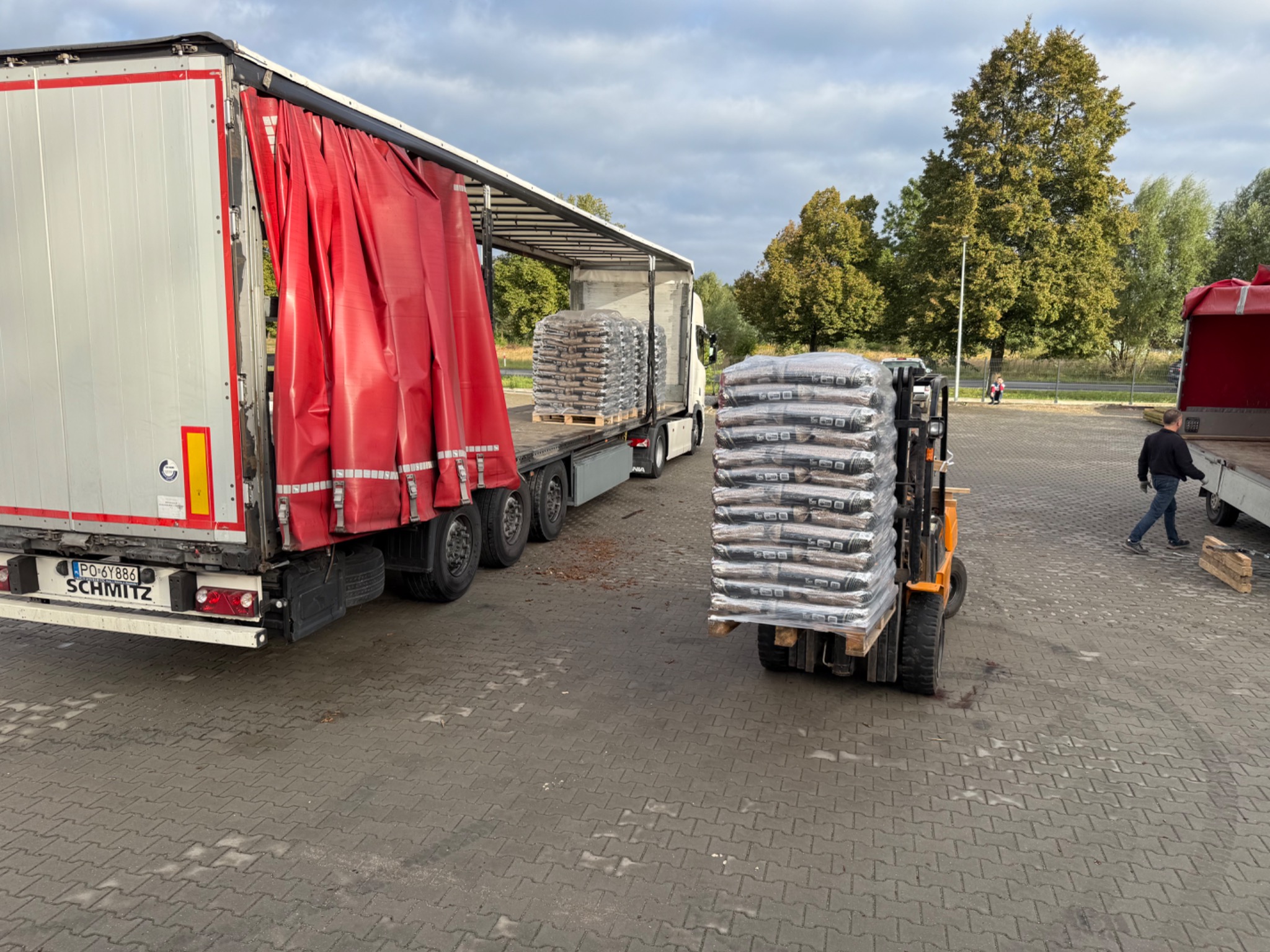 Wózek widłowy przewozi paletę z workami. Częściowo zasłonięta ciężarówka z czerwoną plandeką w tle. Transport pelletu.