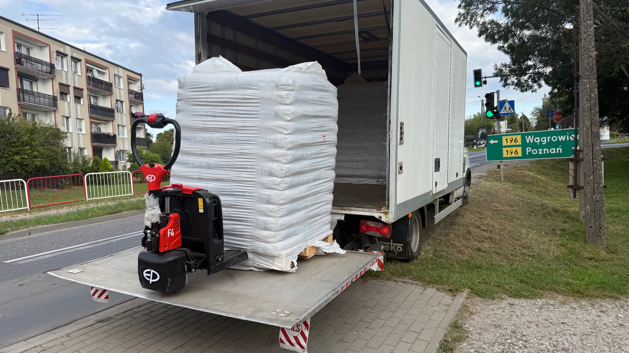 Rozładunek pelletu z ciężarówki za pomocą wózka paletowego, widoczny znak drogowy z kierunkami na Wągrowiec i Poznań. Dzień.