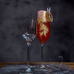 Sławomir Natoński - Kieliszek z czerwonym koktajlem udekorowanym jadalnym złotem i suszoną limonką przypiętą klamerką, obok rozbity kieliszek i fragmenty szkła na ciemnym tle.