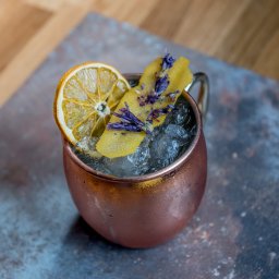 Sławomir Natoński - Miedziany kubek z lodem, udekorowany suszoną pomarańczą, skórką cytryny z lawendą, na tle drewnianego blatu i szarej płyty.