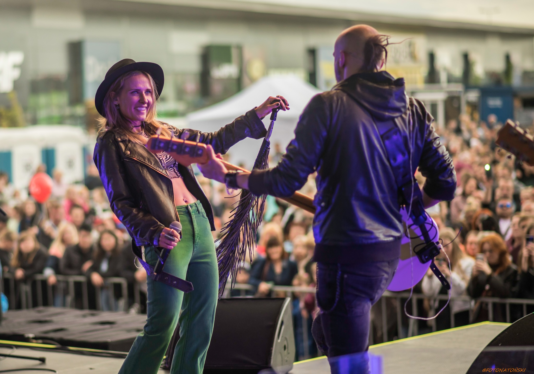 Uśmiechnięta wokalistka w czarnym kapeluszu i skórzanej kurtce, śpiewa na scenie, trzymając mikrofon; w tle gitarzysta i rozmyty tłum publiczności.