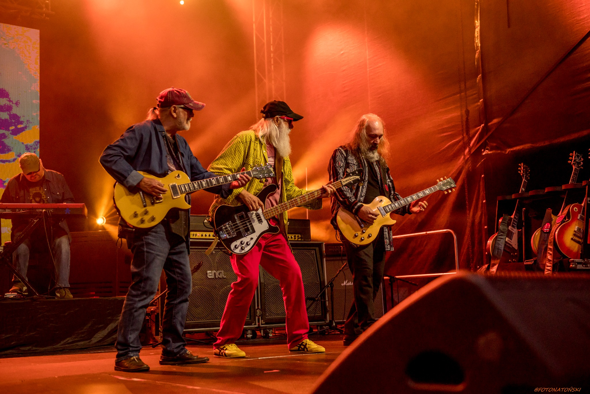 Zespół muzyczny podczas koncertu: trzech gitarzystów z długimi brodami gra na żółtych gitarach, w tle klawiszowiec i rząd gitar na statywach, oświetlenie sceniczne w odcieniach czerwieni.