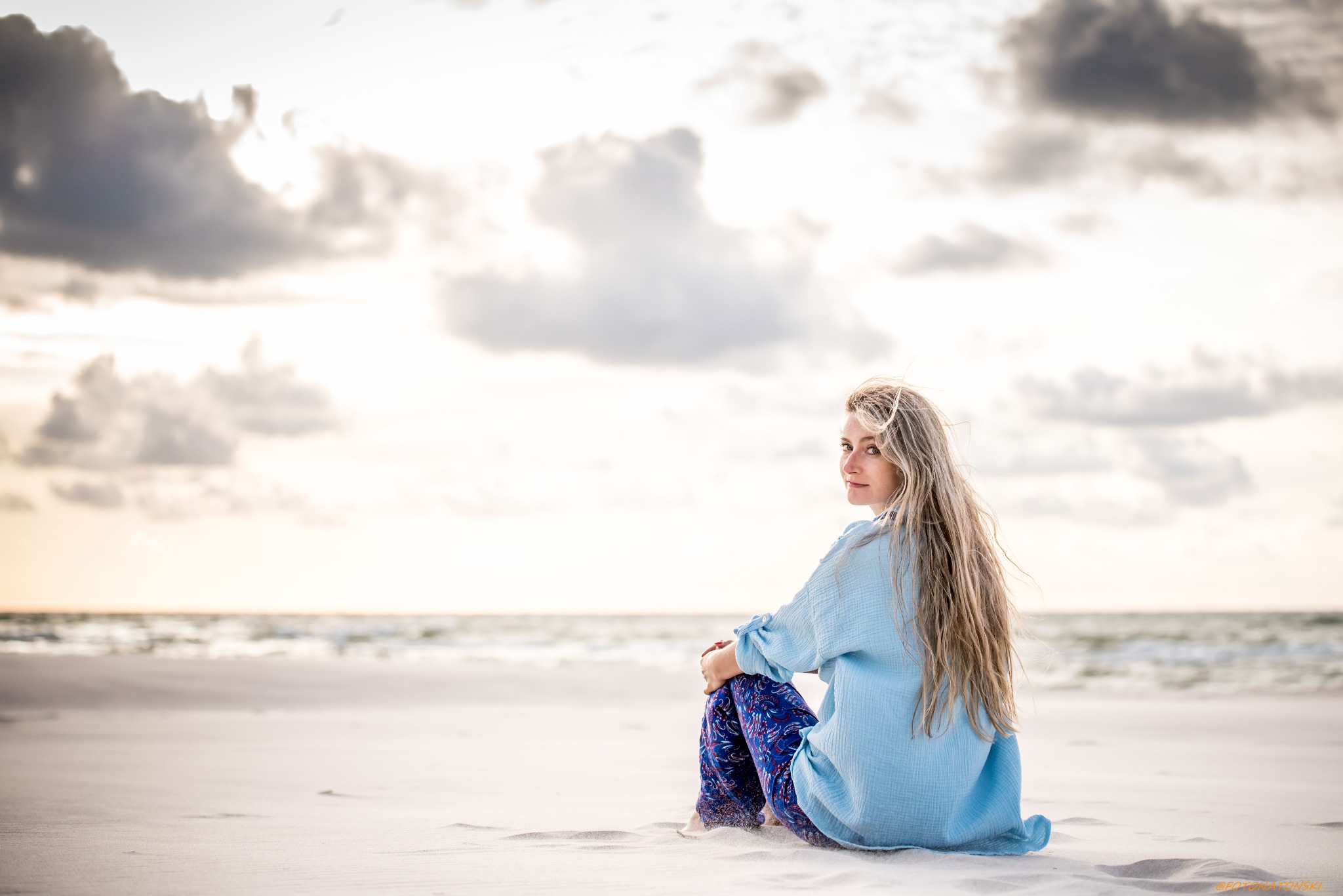 Kobieta z długimi blond włosami siedzi na piaszczystej plaży, ubrana w błękitną bluzkę i wzorzyste spodnie, spoglądając przez ramię w stronę obiektywu, w tle widoczne morze i pochmurne niebo.