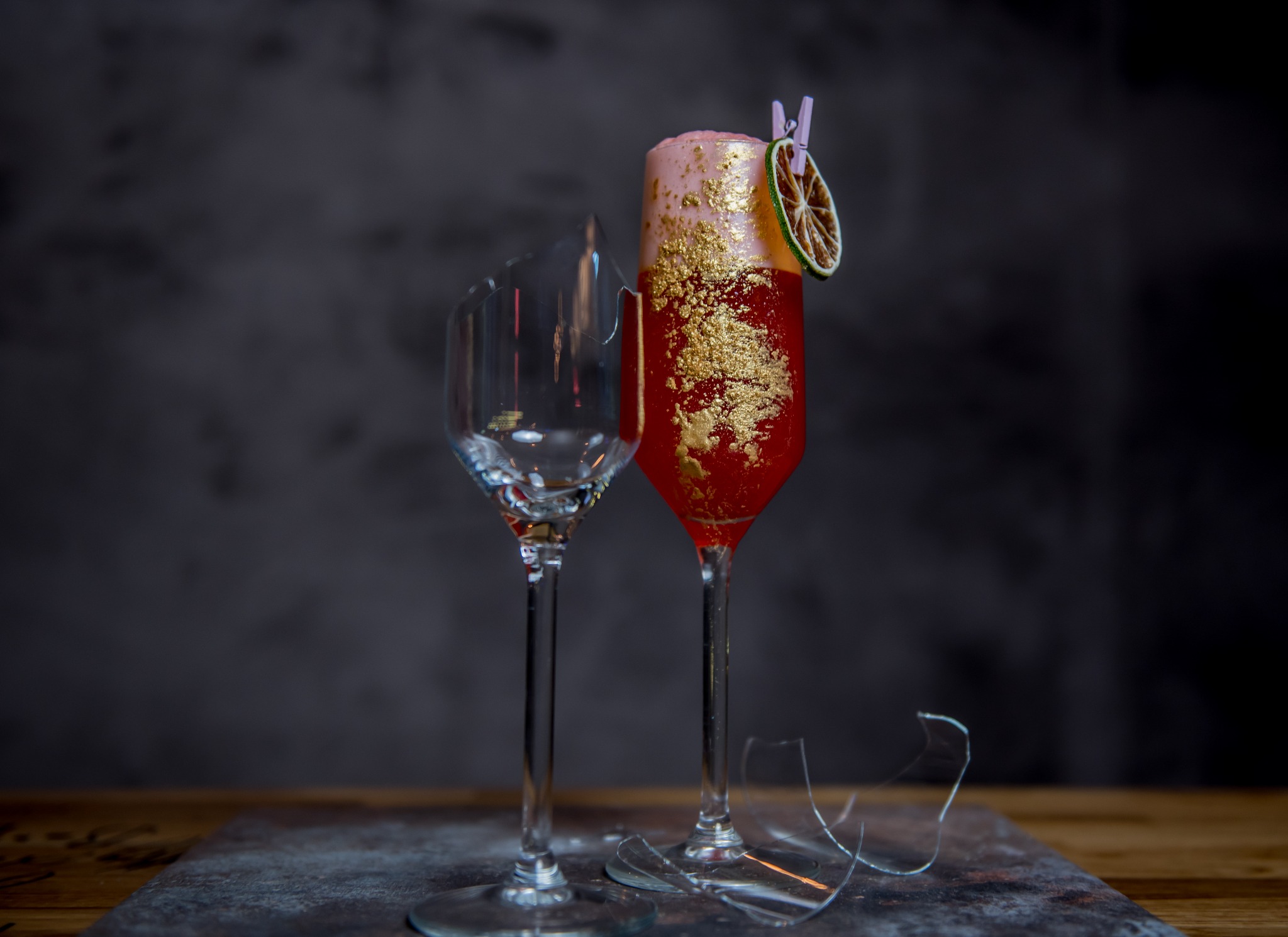 Kieliszek z czerwonym koktajlem udekorowanym jadalnym złotem i suszoną limonką przypiętą klamerką, obok rozbity kieliszek i fragmenty szkła na ciemnym tle.