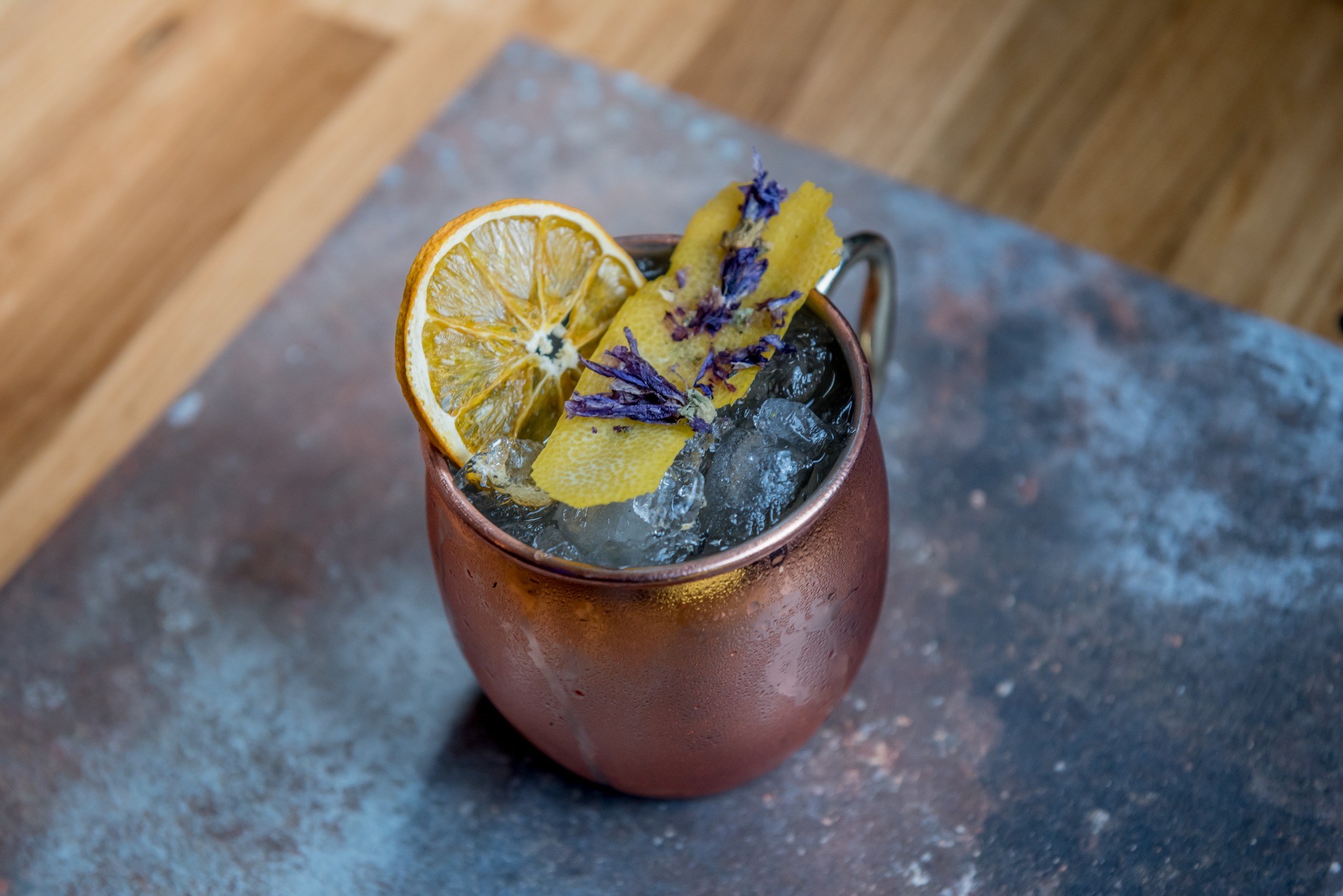 Miedziany kubek z lodem, udekorowany suszoną pomarańczą, skórką cytryny z lawendą, na tle drewnianego blatu i szarej płyty.