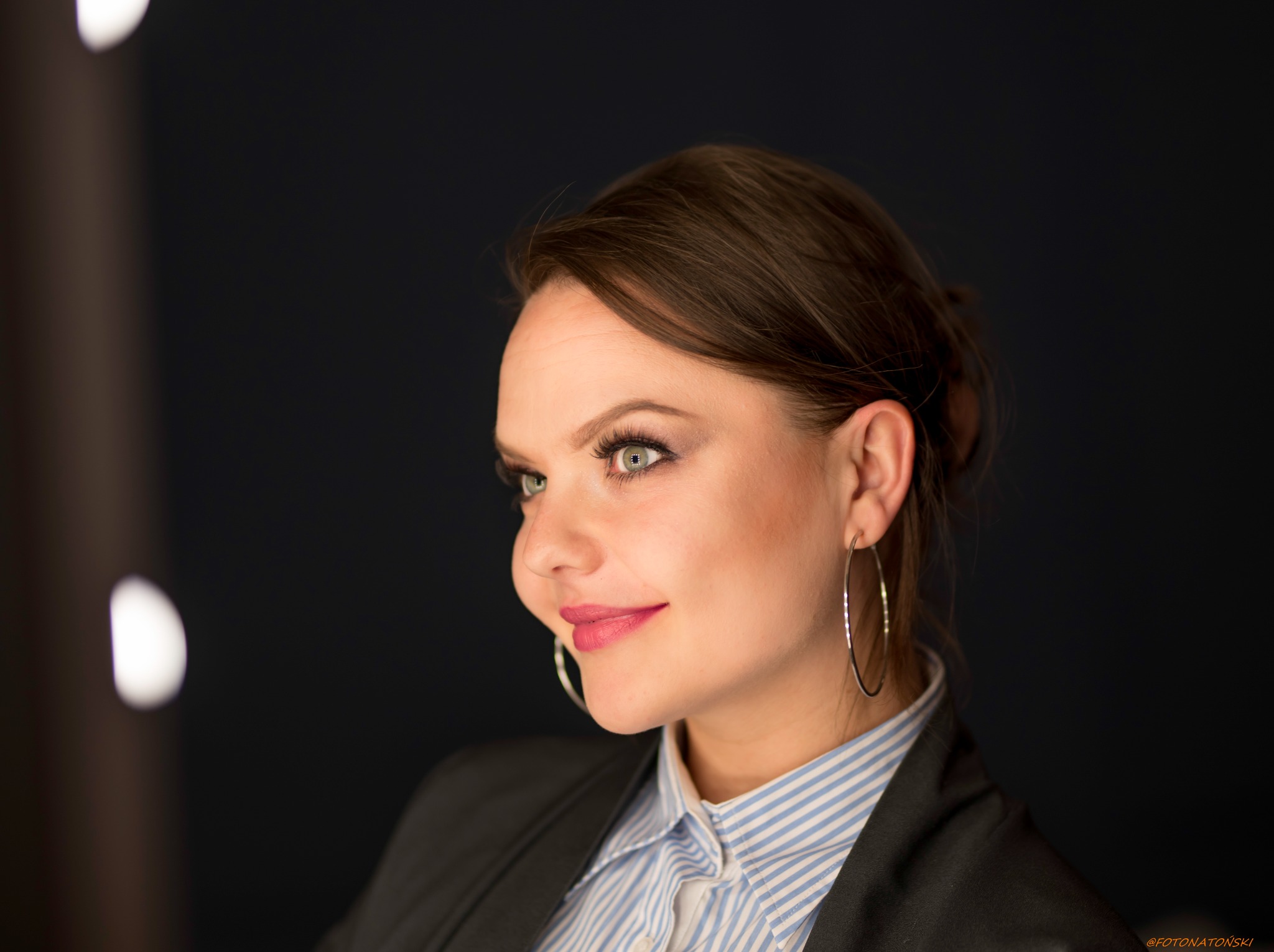 Portret kobiety w koszuli w paski i marynarce, z kolczykami kołami, patrzy w lewo, z delikatnym uśmiechem i ciemnym tłem.