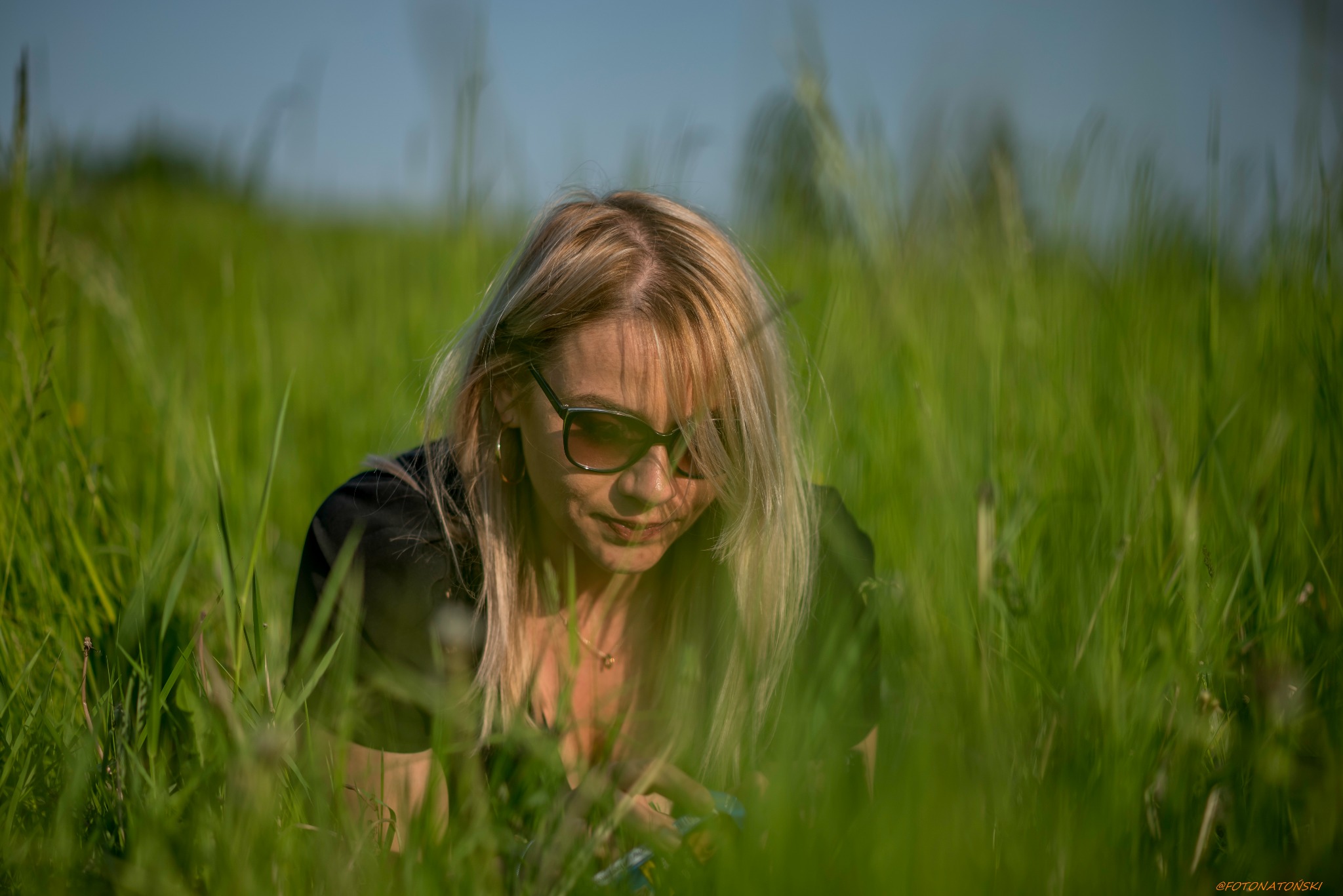 Portret kobiety z blond włosami i okularami przeciwsłonecznymi leżącej w wysokiej trawie, patrzącej w dół, z delikatnym uśmiechem.