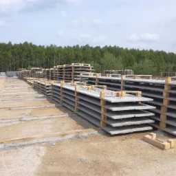Stosy prefabrykowanych płyt betonowych z wystającymi elementami stalowymi, składowane na placu budowy, w tle widoczny las.