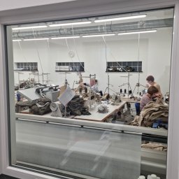 Militarjach Grzegorz Jach - Widok przez okno na dużą, jasną halę z kilkoma stanowiskami pracy z maszynami do szycia, przy których pracują osoby szyjące odzież z materiałów o różnych odcieniach brązu i zieleni.