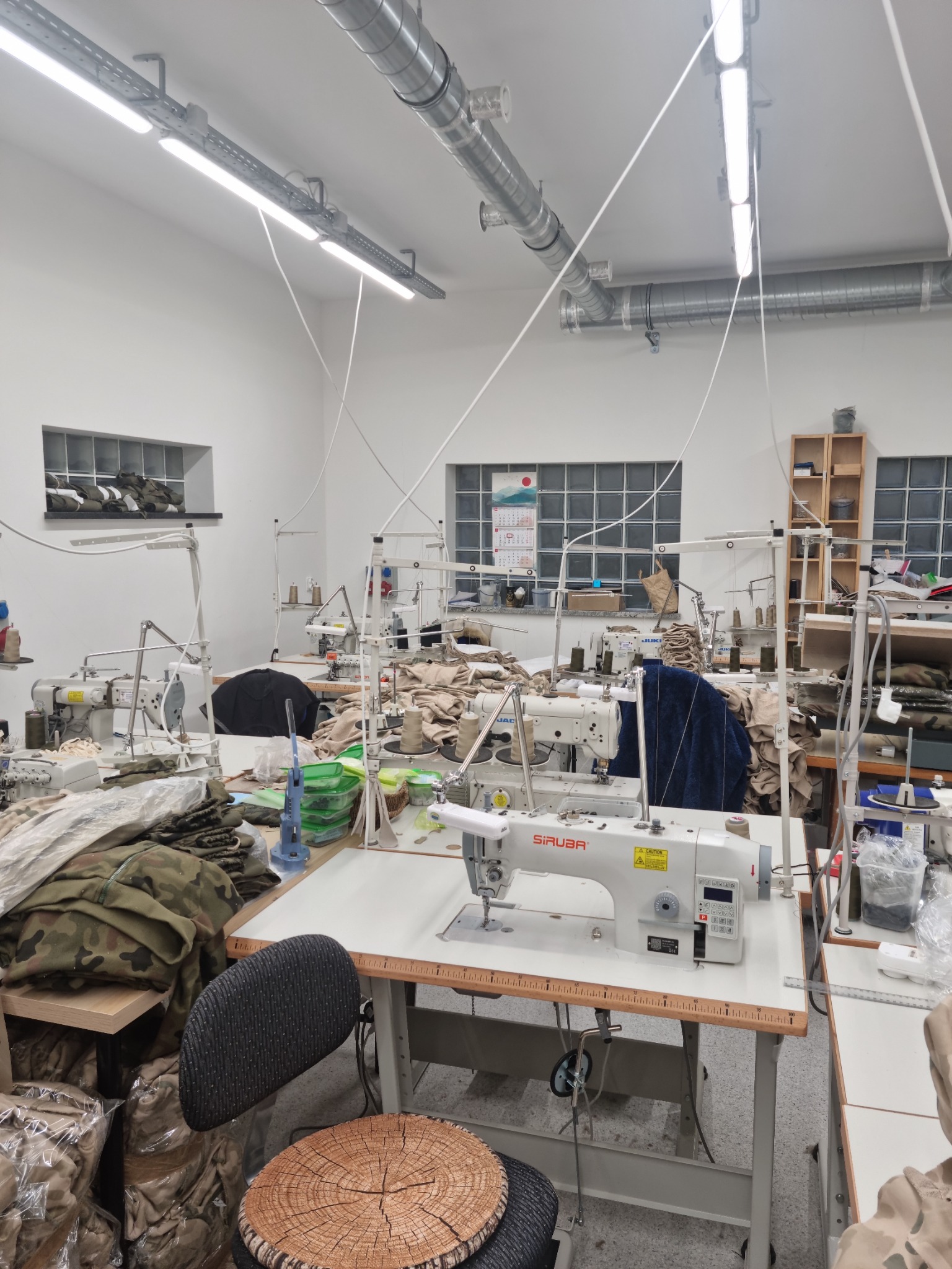 Jasne wnętrze pracowni krawieckiej z kilkoma stanowiskami wyposażonymi w maszyny do szycia Siruba, stosy materiałów w kamuflażu wojskowym i krzesło z siedziskiem imitującym plaster drewna.
