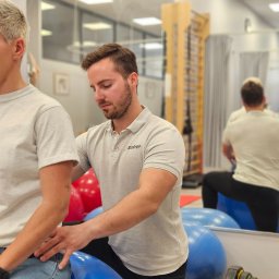 Salveo Medical Care Katowice Sp. z o.o - Mężczyzna w szarej koszulce pomaga pacjentce siedzącej na niebieskiej piłce gimnastycznej w sali rehabilitacyjnej z drabinkami i lustrem.