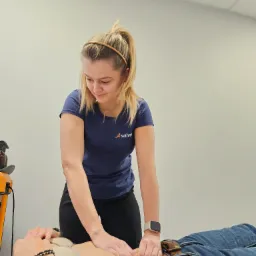 Kobieta w niebieskiej koszulce uciska dłońmi brzuch pacjenta leżącego na plecach, w tle biała ściana i sprzęt medyczny, widoczne detale: zegarek na ręku, biżuteria.