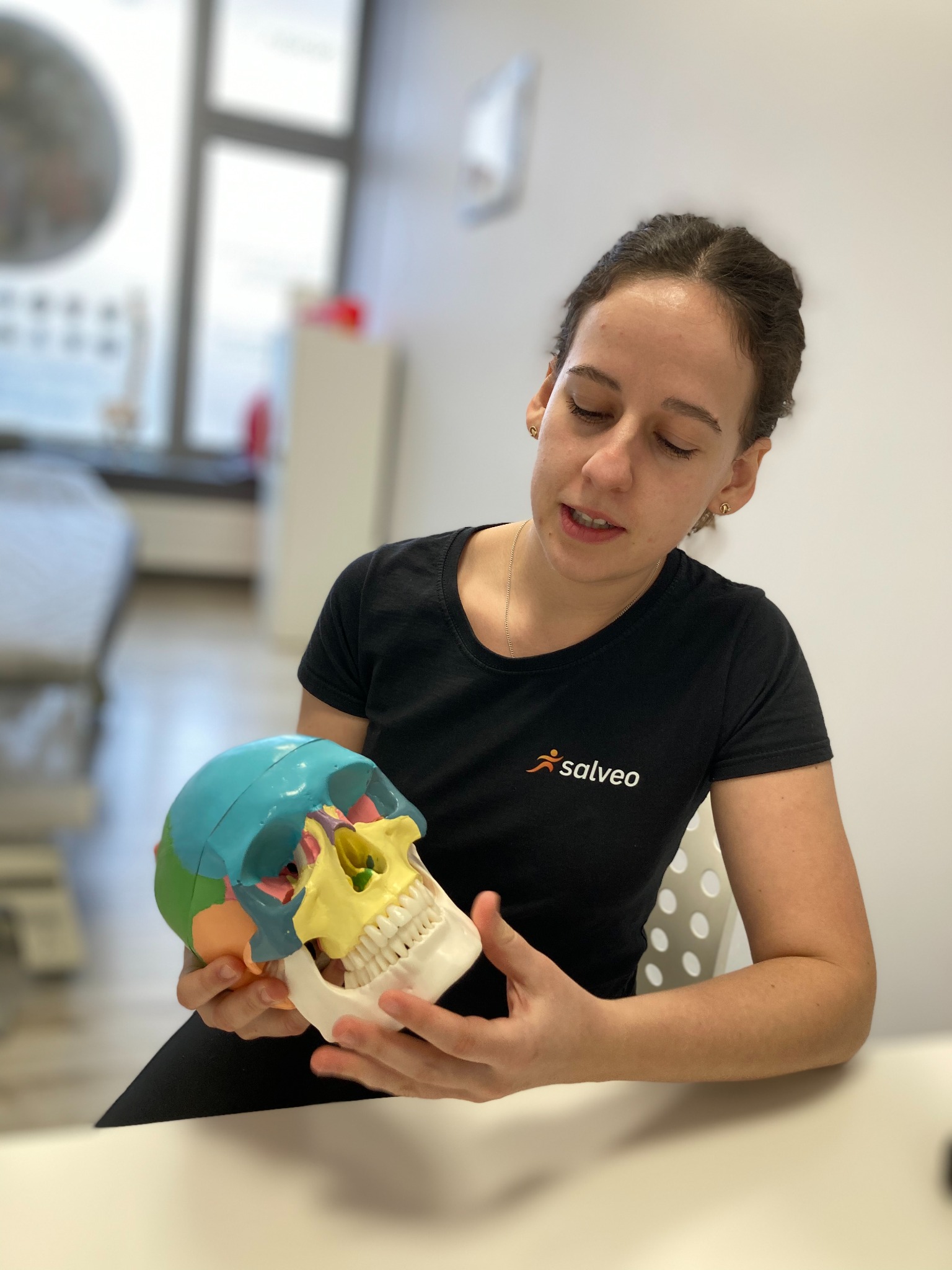 Kobieta w czarnej koszulce z logo Salveo trzyma kolorowy model czaszki, demonstrując anatomię. Jasne wnętrze gabinetu, naturalne światło, skupienie na edukacji.