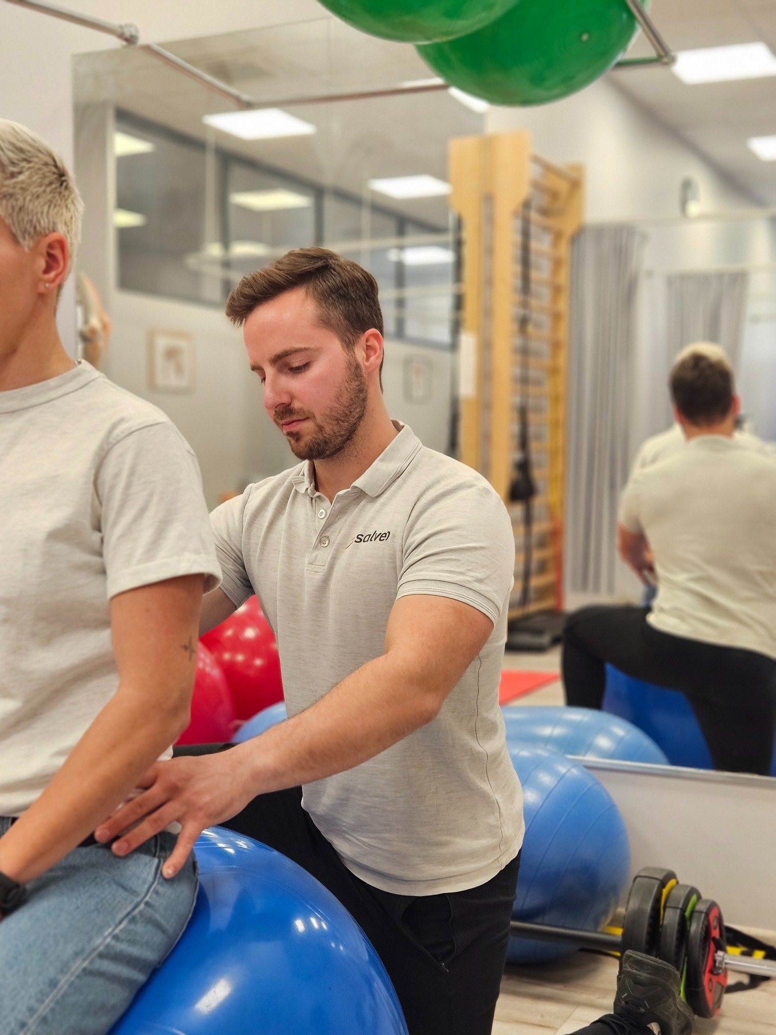 Mężczyzna w szarej koszulce pomaga pacjentce siedzącej na niebieskiej piłce gimnastycznej w sali rehabilitacyjnej z drabinkami i lustrem.