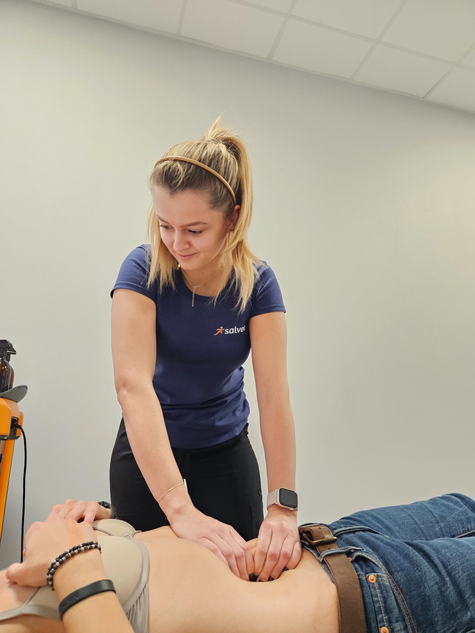 Kobieta w niebieskiej koszulce uciska dłońmi brzuch pacjenta leżącego na plecach, w tle biała ściana i sprzęt medyczny, widoczne detale: zegarek na ręku, biżuteria.