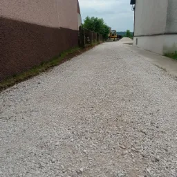 Droga gruntowa wysypana kruszywem, widoczna koparka w oddali, pomiędzy dwoma budynkami, pochmurne niebo.