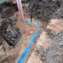 Wykop z piaskowym podłożem, w którym ułożono niebieską rurę w folii ochronnej, widoczna instalacja pionowa w osłonie, ziemia z kamieniami i fragment ogrodzenia z siatki w tle.