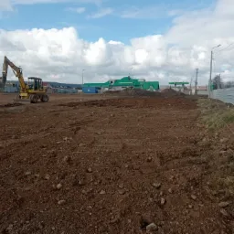 Żółta koparka kołowa na placu budowy, przygotowująca teren pod inwestycję; w tle widoczny budynek stacji benzynowej i błękitne niebo z chmurami.