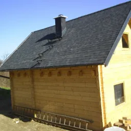 Drewniany domek z ciemnoszarym dachem pokrytym gontem bitumicznym, widoczne zabezpieczenia przeciwśniegowe wzdłuż dachu, komin i rynna. Drabina oparta o ścianę budynku.