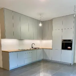 Jasnoszara zabudowa kuchenna z drewnianym blatem, czarną armaturą i płytą indukcyjną, oświetlenie podszafkowe, szare płytki podłogowe, surowa żarówka zwisająca z sufitu.