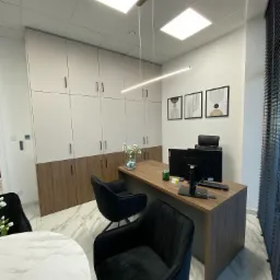 Nowoczesne biuro z drewnianym biurkiem, czarnymi krzesłami welurowymi, białymi szafkami z drewnianymi akcentami, minimalistycznymi obrazami na ścianie i marmurową podłogą.