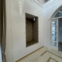 Biała szafa wnękowa z tapicerowanym siedziskiem w Jasienicy, oświetlonym kinkietem, kryształowy żyrandol odbija się w lakierowanych frontach.