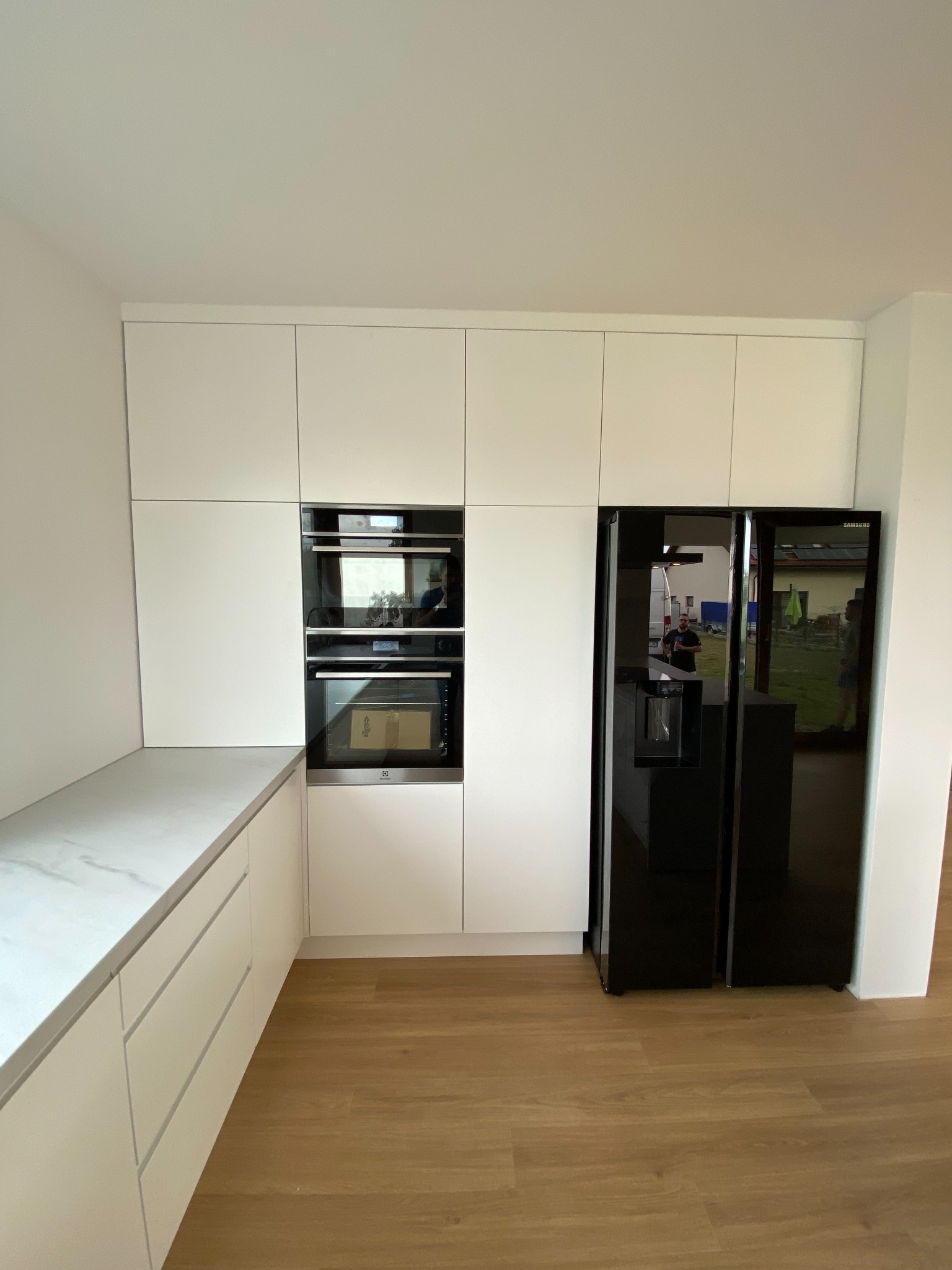 Minimalistyczna, biała zabudowa kuchenna z wbudowanym piekarnikiem i czarną lodówką side-by-side, widoczne odbicie osoby w drzwiach lodówki.