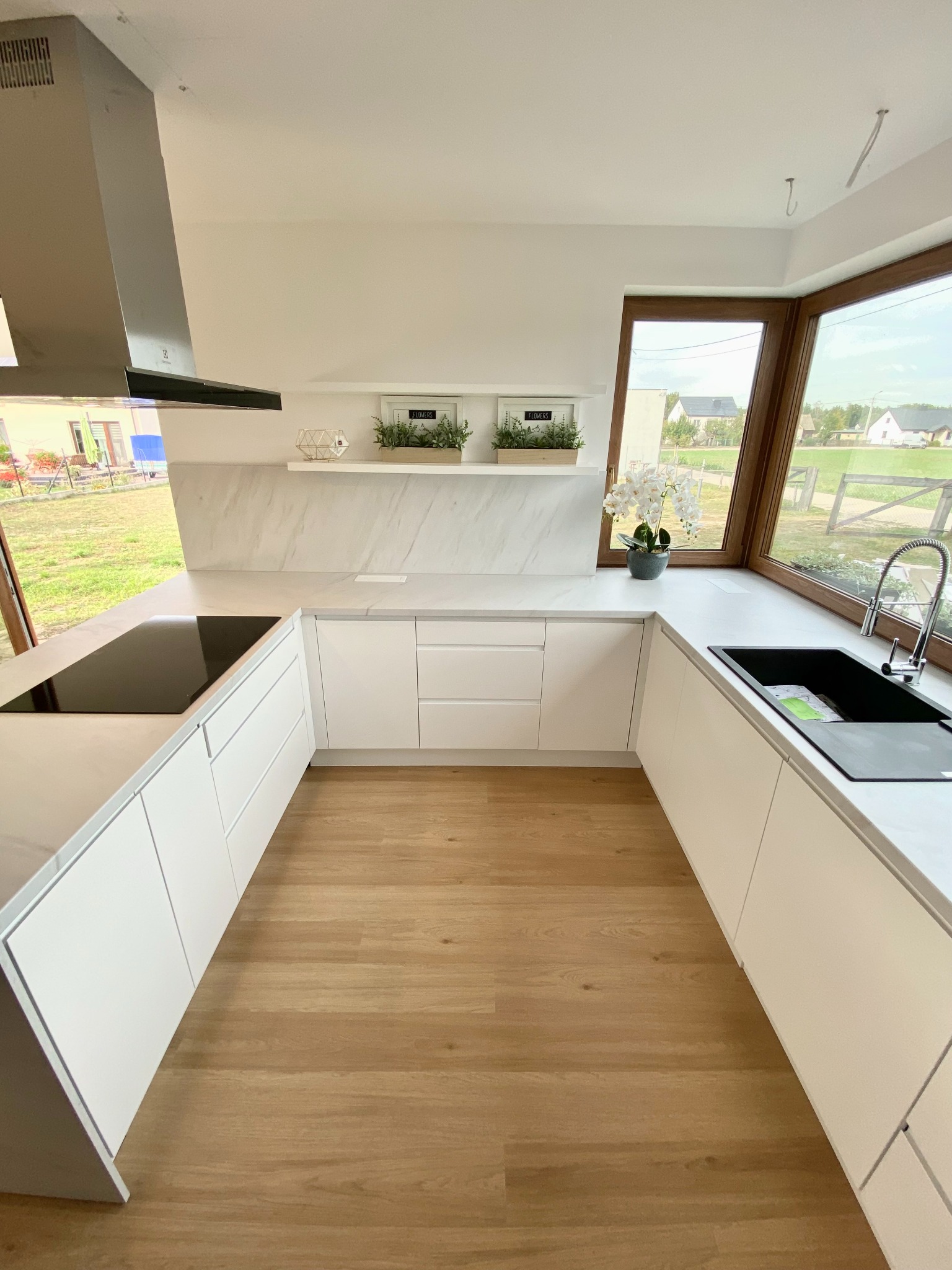 Jasna kuchnia z białymi szafkami, marmurowym blatem i drewnianą podłogą, widok na ogród przez duże okno.