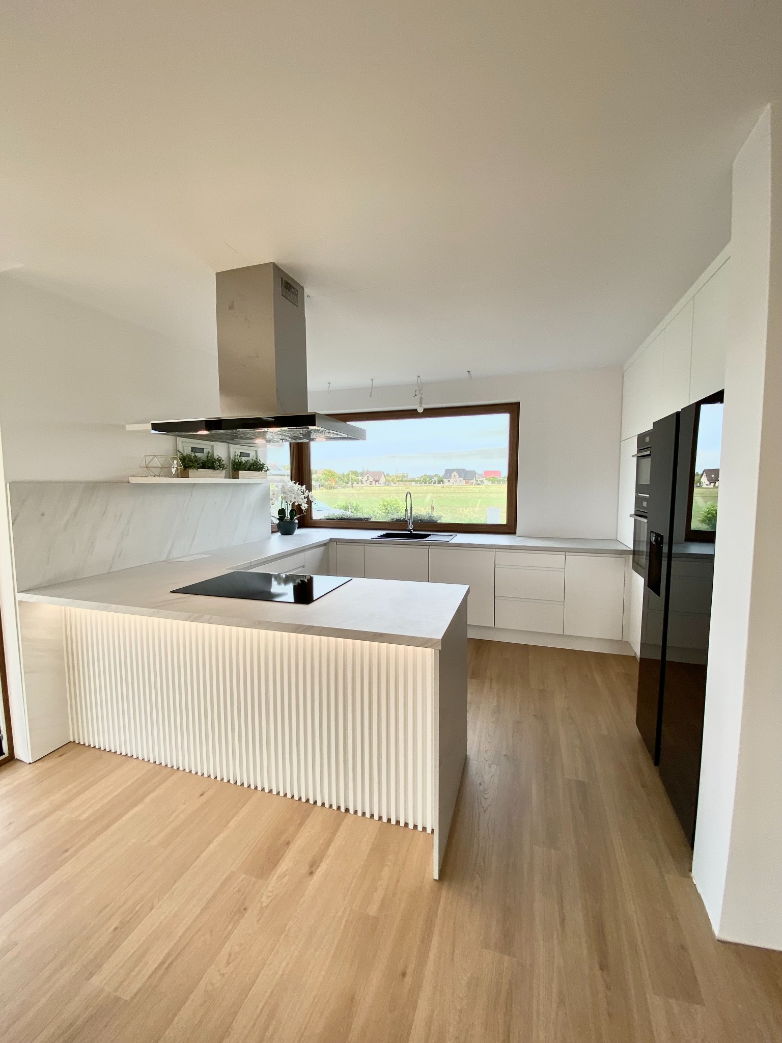 Jasna kuchnia z wyspą oświetloną od dołu, płyta indukcyjna i okap nad wyspą, widok na zielone pola za oknem.