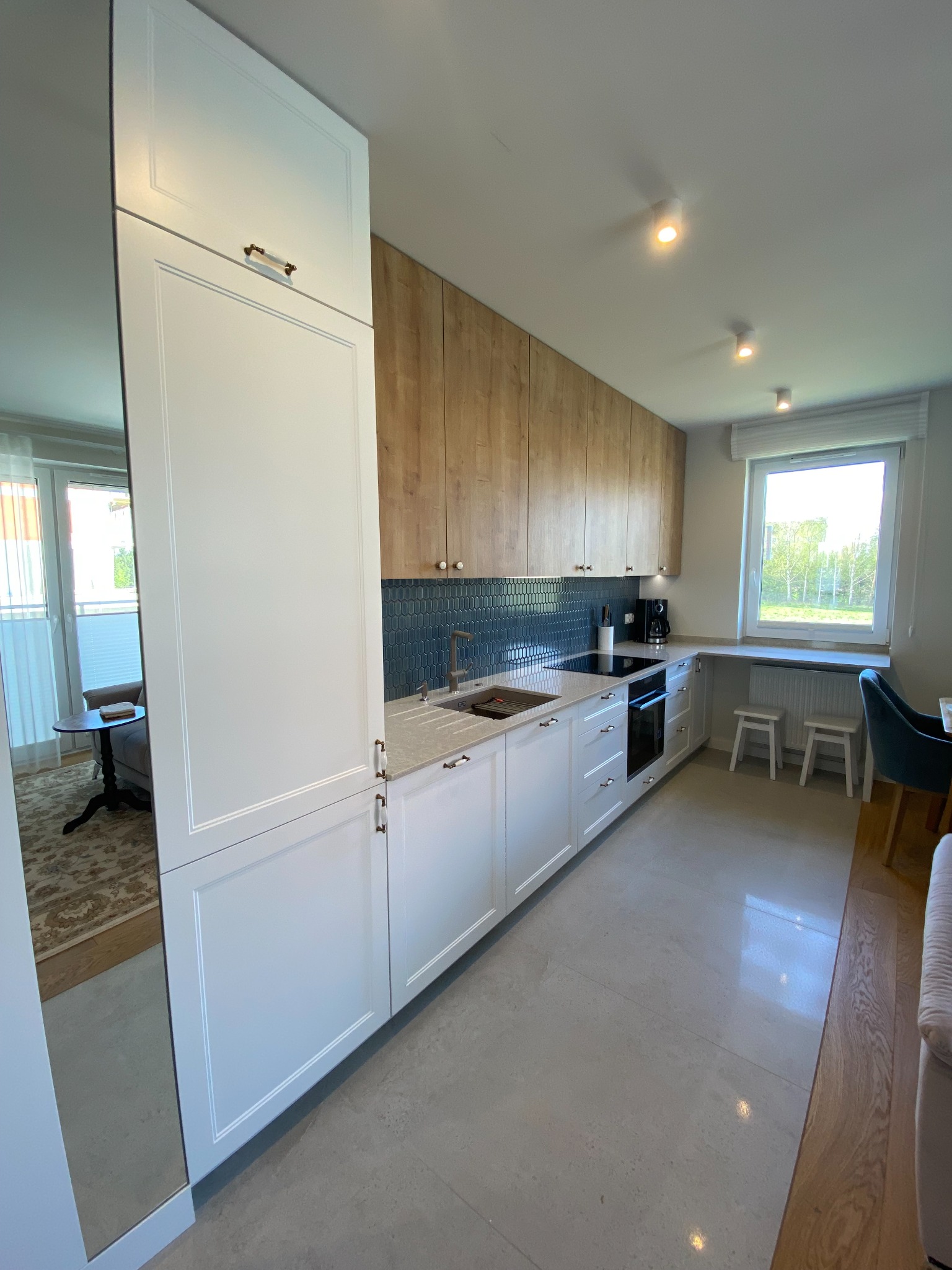 Nowoczesna kuchnia z białymi szafkami dolnymi i drewnianymi górnymi, niebieska mozaika nad blatem, widok na salon przez lustrzane drzwi.