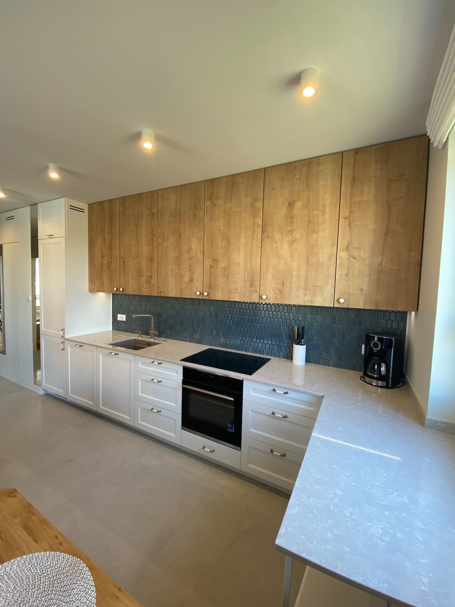 Jasna kuchnia z drewnianymi szafkami górnymi i białymi dolnymi, ciemnoniebieska mozaika na ścianie, ekspres do kawy na blacie.