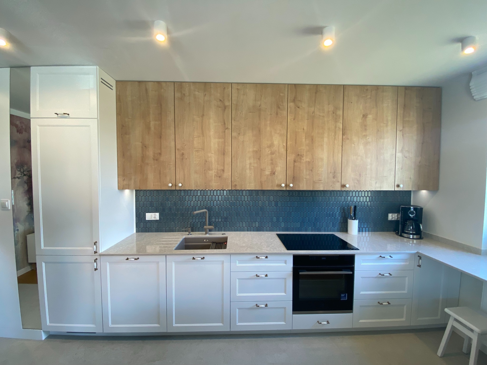 Nowoczesna kuchnia z białymi dolnymi szafkami i drewnopodobnymi górnymi, niebieska mozaika na ścianie, blat z jasnego kamienia.