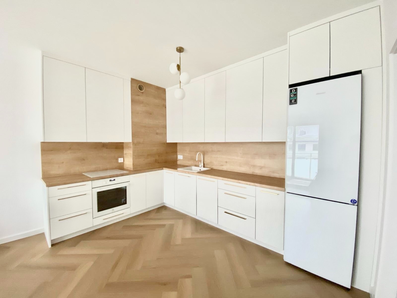 Jasna kuchnia z białymi szafkami i drewnianymi blatami, parkiet ułożony w jodełkę, nowoczesna lampa wisząca z trzema kulami.