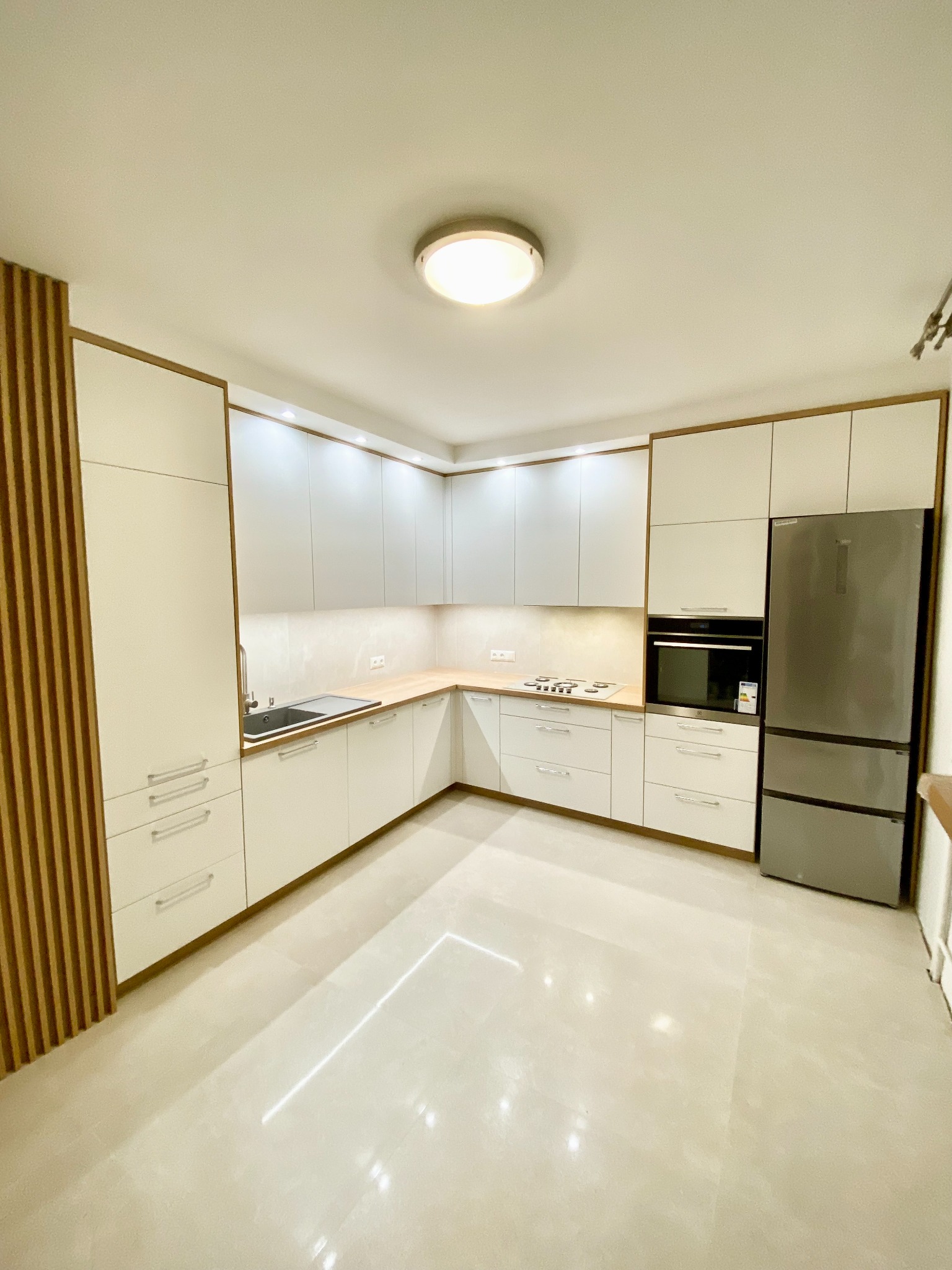 Nowoczesna kuchnia w jasnych barwach z drewnianymi akcentami, widok na narożną zabudowę z białymi frontami, drewnianym blatem, stalowym zlewem i lodówką.