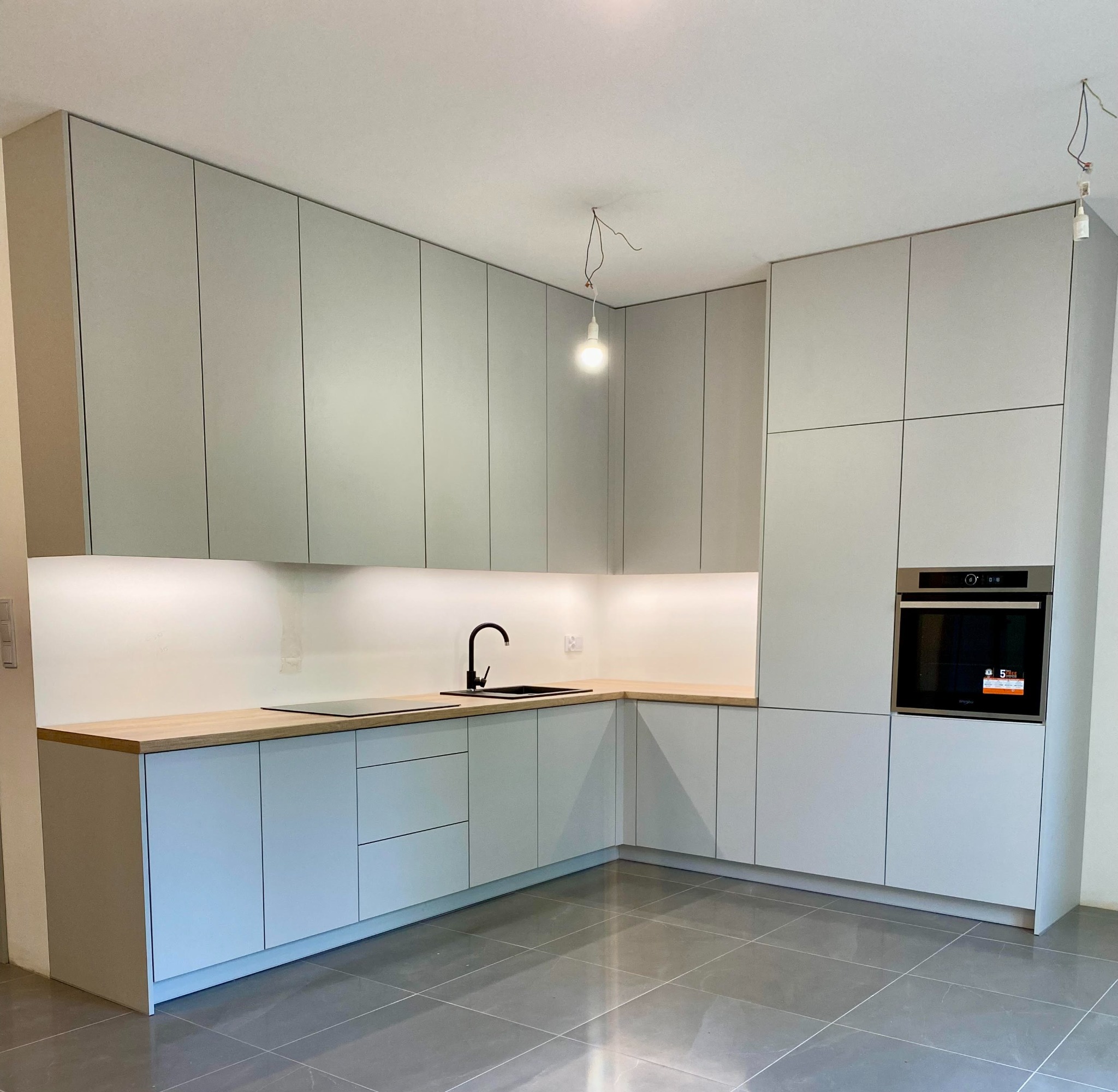 Jasnoszara zabudowa kuchenna z drewnianym blatem, czarną armaturą i płytą indukcyjną, oświetlenie podszafkowe, szare płytki podłogowe, surowa żarówka zwisająca z sufitu.