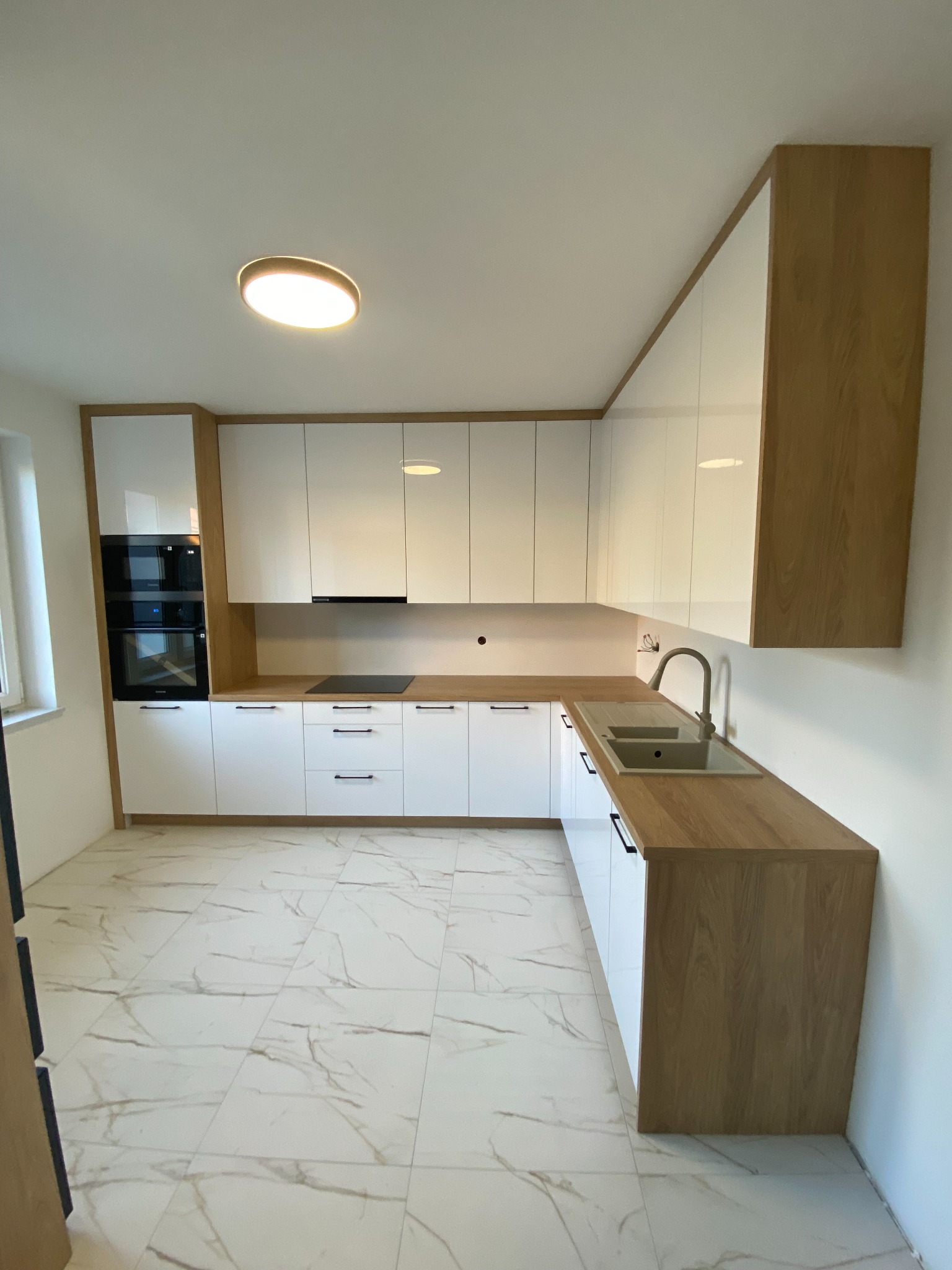 Nowoczesna kuchnia z białymi, błyszczącymi frontami i drewnianymi blatami, widok na narożną zabudowę z piekarnikiem i płytą indukcyjną, marmurowe płytki na podłodze, okrągła lampa sufitowa.